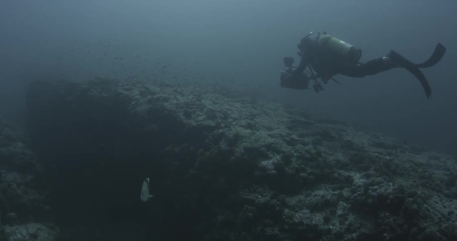 A diver with a camera is being filmed from the side swimming towards a large opening