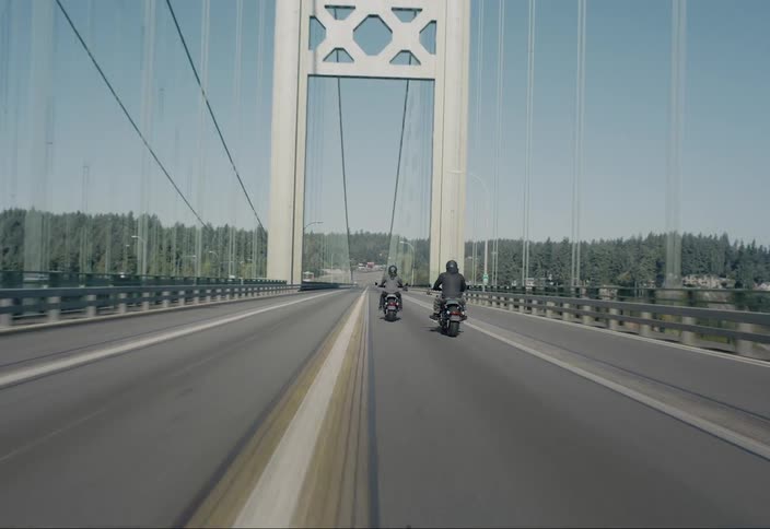 Two motorcyclists riding along a highway passing houses surrounded by forest cross a bridge over the water during the day