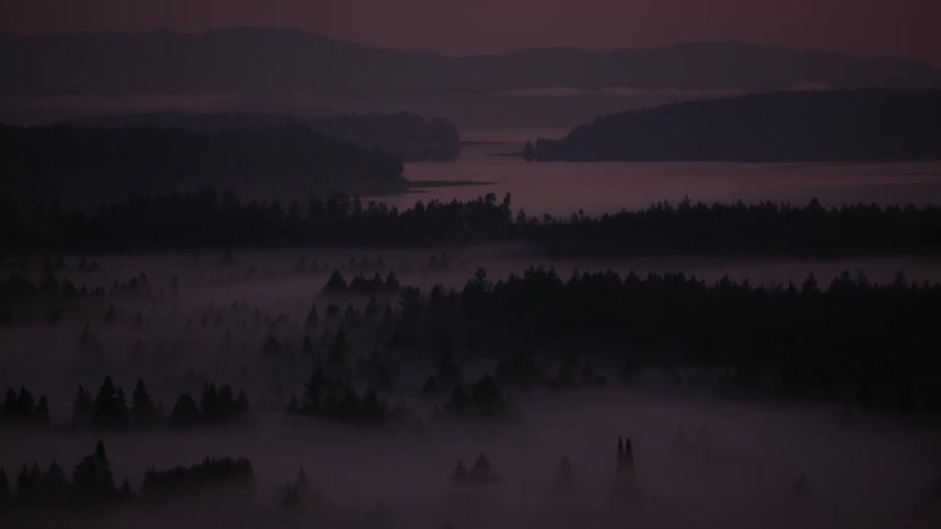 A hazy aerial view shows a forest and hills shrouded in fog overlooking a calm lake at twilightdawn