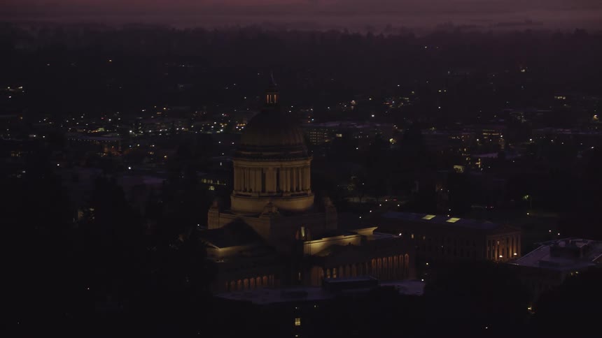 At dusk the illuminated dome of the washington state capitol building stands out against the dark silhouette of the surrounding city and landscape