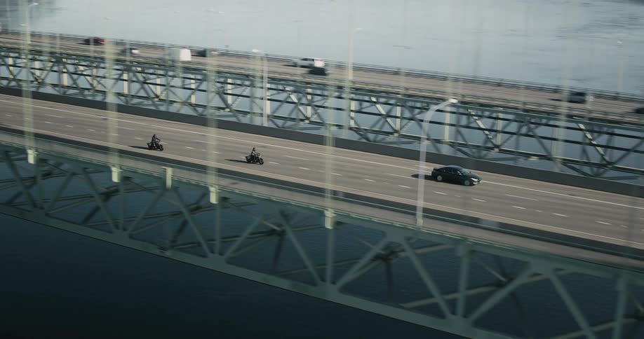 Motorcyclists ride across a busy bridge between forested banks and along a highway passing a town
