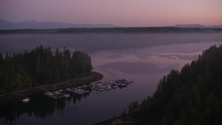 A tranquil bay shrouded in early morning mist is surrounded by densely forested hills with a few boats moored in the calm water