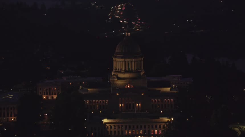 Neoclassical building with dome illuminated at night surrounded by other structures and scattered points of light