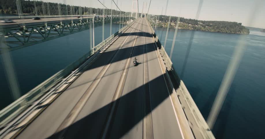 A lone motorcycle rider travels down the center of a long modern bridge spanning a wide blue body of water with forested shores visible in the distance