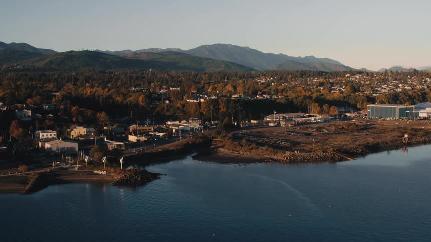 An aerial view flies up revealing a coastal town situated beside a bay with distant hills and mountains