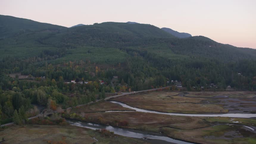An aerial view reveals a rural town nestled in a valley with a river fields and surrounding forested hills