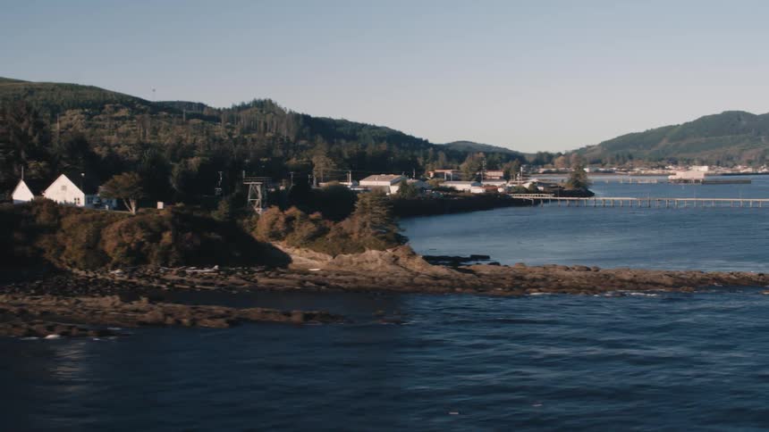A rocky point extends into a bay with buildings nestled among trees and a forested hill rising in the background