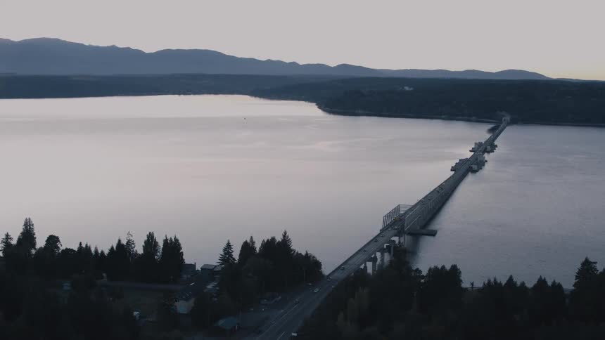 An aerial view shows a long bridge over a lake with cars on it and boats passing underneath