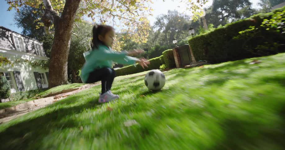 Young girl crouches to catch a soccer ball in a residential yard under a larger tree with its autumn foliage