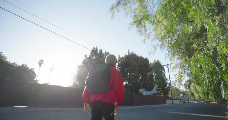 A teenager in a red cap and jacket is skateboarding on an asphalt street and he is wearing a backpack on his back