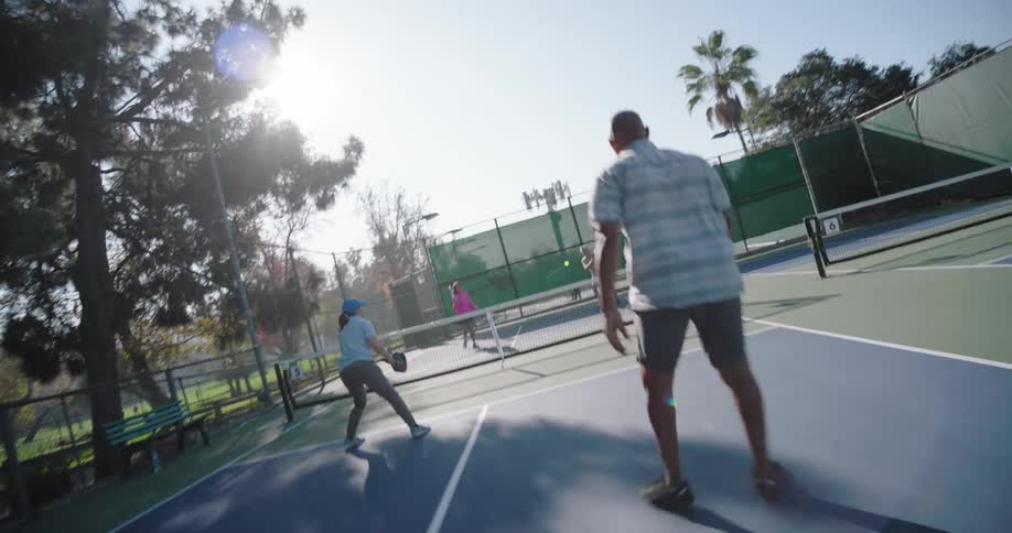 Two couples playing pickleball on an outdoor court surrounded by tall trees and a fence