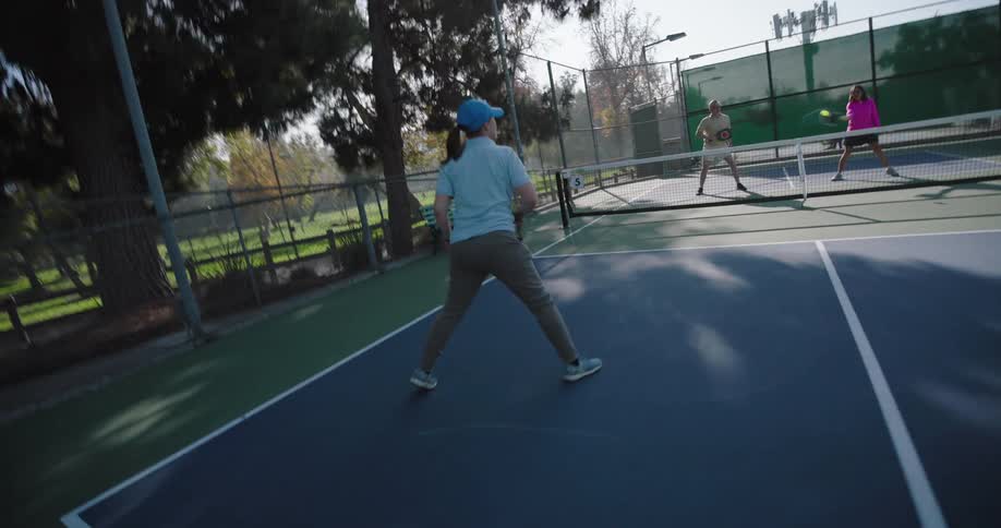 Two couples playing pickleball on an outdoor court surrounded by tall trees and a fence