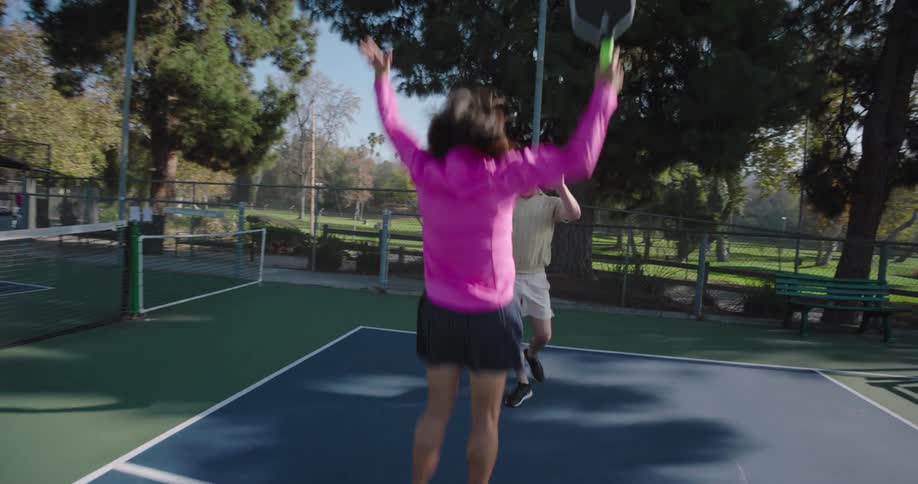 A couple celebrates after winning a game of pickleball on an outdoor court surrounded by tall trees and a fence