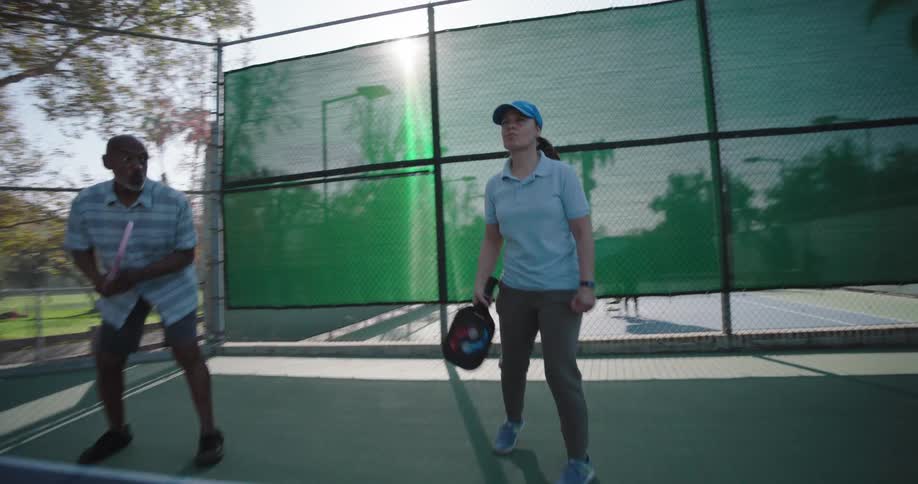 A couple playing pickleball on an outdoor court surrounded by tall trees and a fence