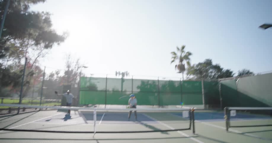 Two couples playing pickleball on an outdoor court surrounded by tall trees and a fence