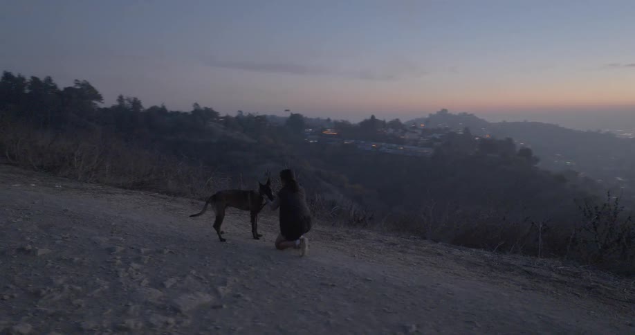 A person is kneeling on a dirt path at dusk holding the leash of a dog with a cityscape and hills visible in the background