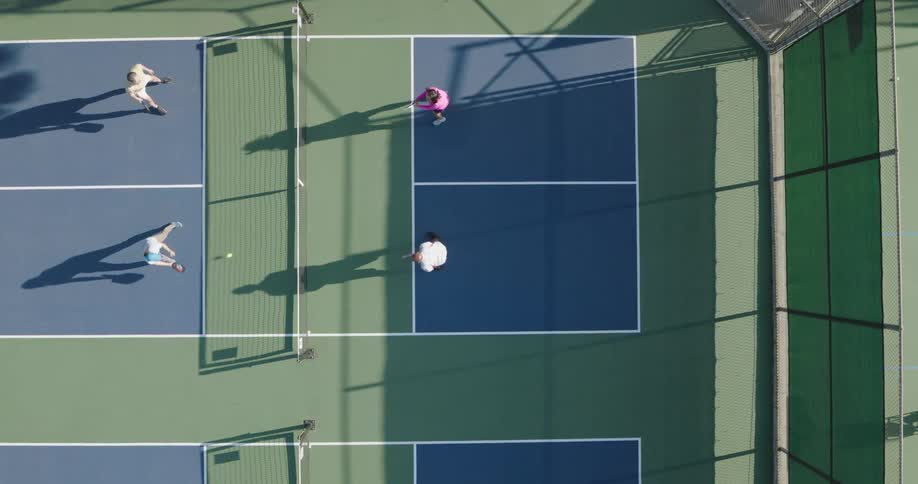 An overhead view of a pickleball court with four players engaged in a game casting long shadows on the court surface