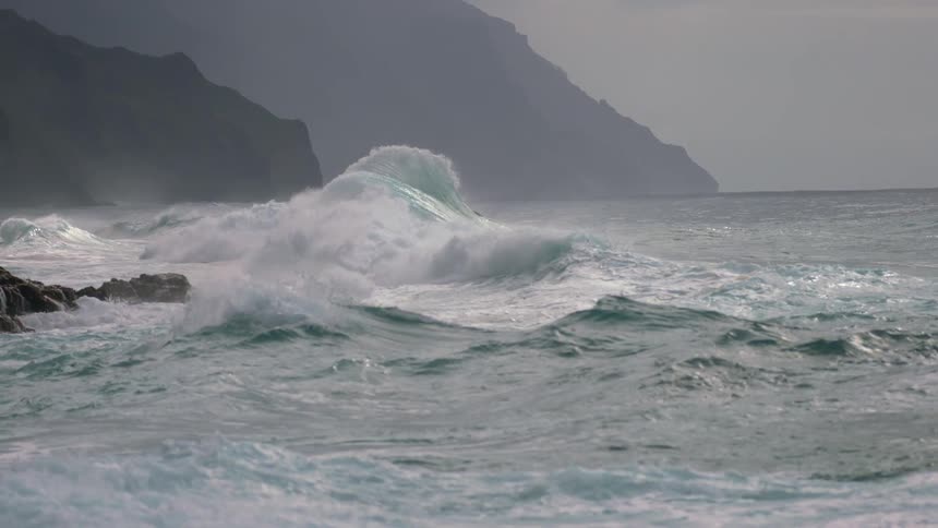 A large wave crashes against a rocky shore sending white spray into the air under an overcast sky with distant mountains in the background