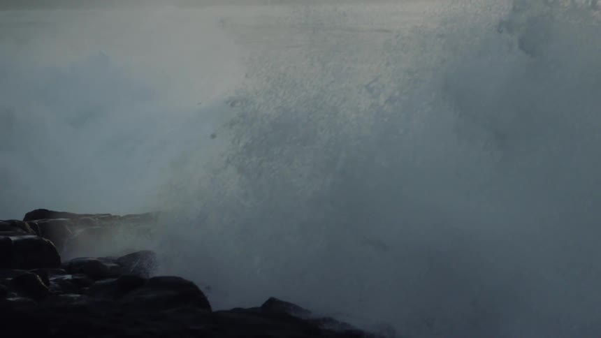 Waves crash against jagged rocks in a misty gray seascape shrouded in a thick fog