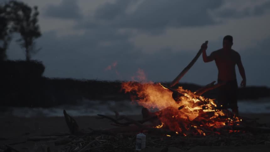 A person is standing on a beach at dusk throwing sticks to a large bonfire