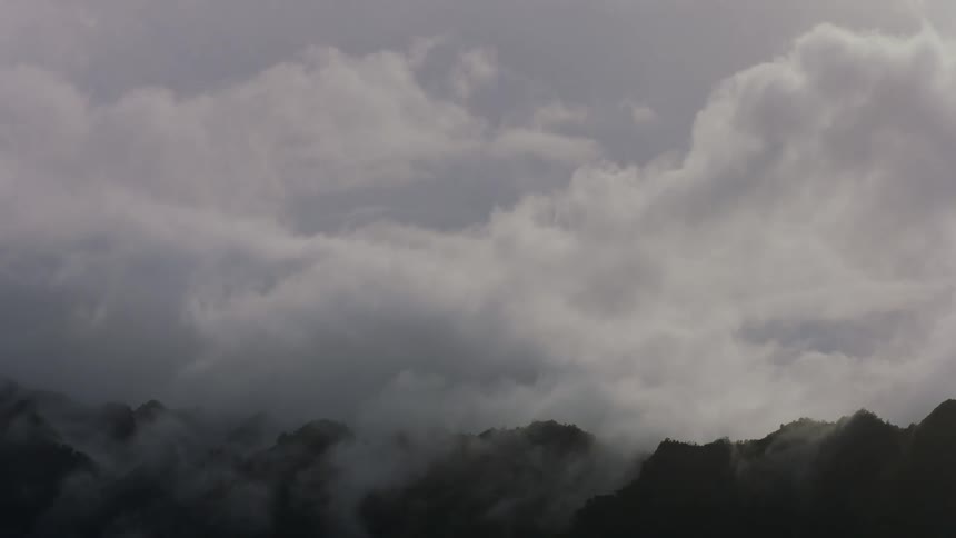 Silhouetted mountain peaks emerge from a swirling mass of lowhanging clouds under a gray sky