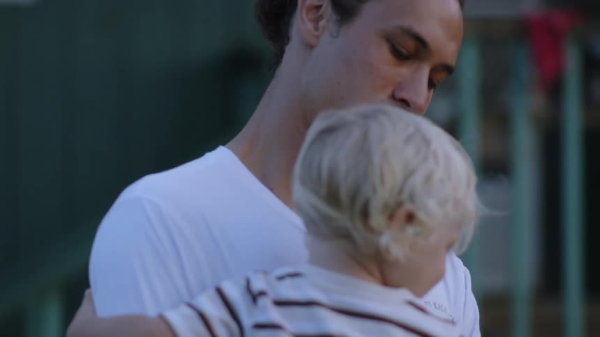 A closeup shows a man in a white shirt holding a child with blond hair wearing a striped shirt in a hug