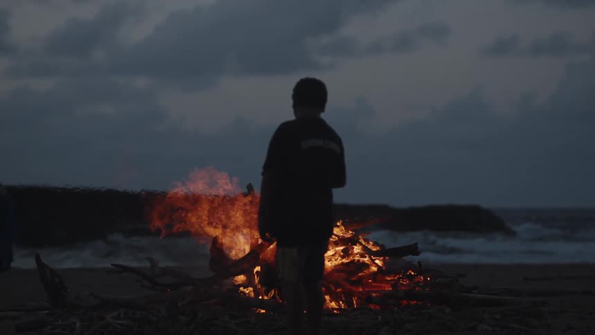 Silhouette of a person standing in front of a bonfire at night on the beach