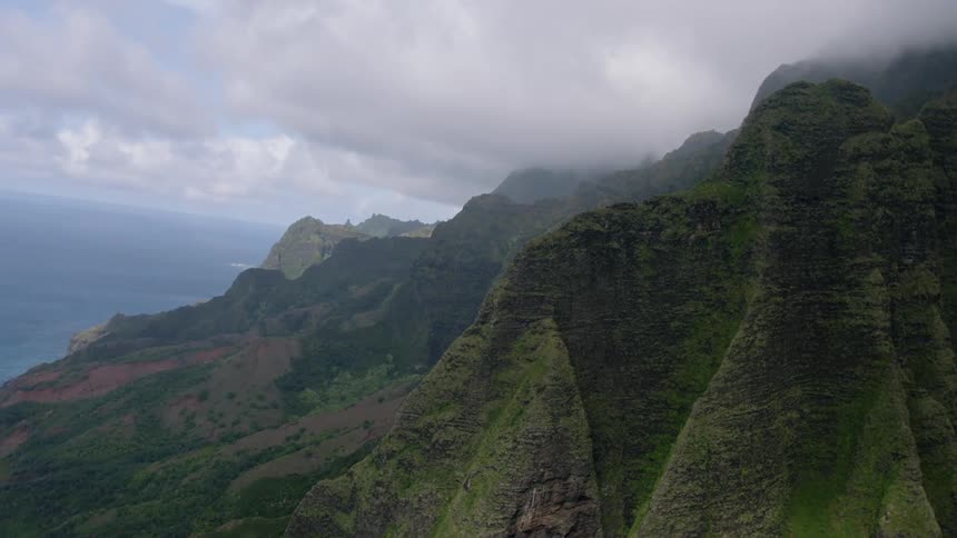 Jagged verdant cliffs rise dramatically from the ocean under a cloudy sky their peaks shrouded in mist