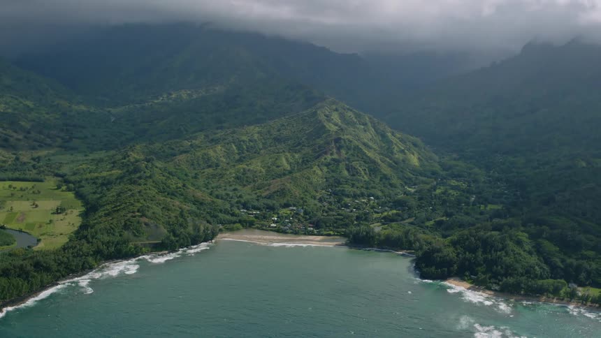 A secluded bay with a sandy beach is framed by lush green mountains under a cloudy sky
