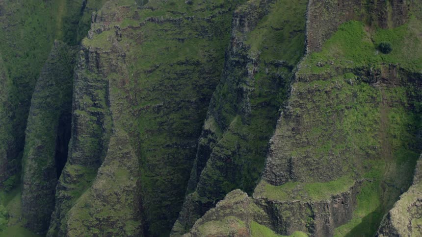 A panningup aerial view showcases steep green and textured cliffs and mountain slopes