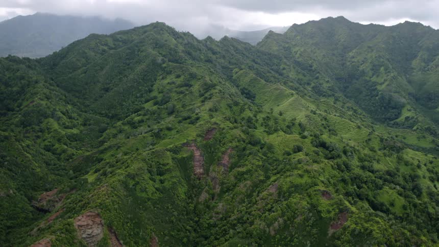 Multiple jagged dark green mountain ridges marked with rocky outcroppings and patches of vegetation extend into the distance under a cloudy overcast sky