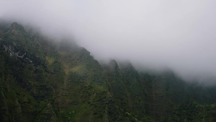 Jagged green cliffs rise steeply their peaks disappearing into a thick lowhanging fog that obscures the upper portions of the rugged terrain