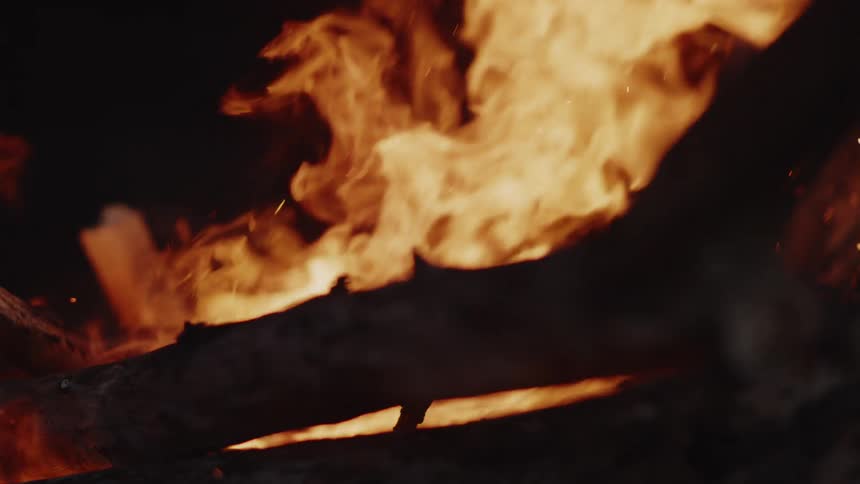 Bright orange flames engulf dark logs with embers glowing and floating upwards against a dark background