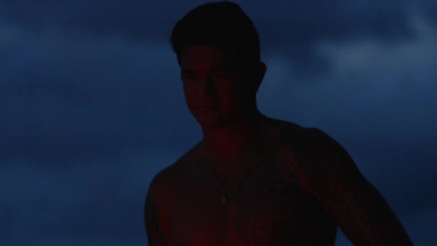 A shirtless muscular man illuminated by the glow of a bonfire at night