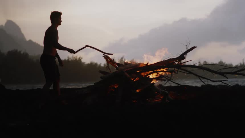 Man on a beach placing a dry branch on a burning fire