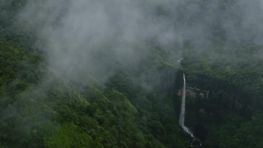 An aerial view of a waterfall in the middle of a green forest with a fog in the background
