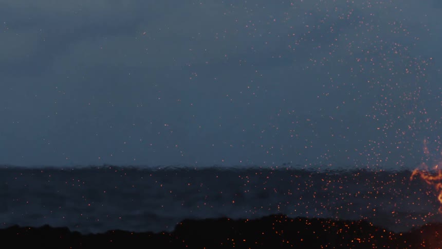 A bonfire burns against a dark night sky sending sparks upward in front of the ocean