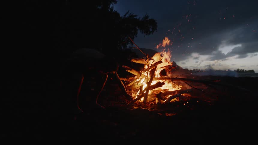 At night a silhouetted person tends to a bright campfire on a beach with waves crashing in the background