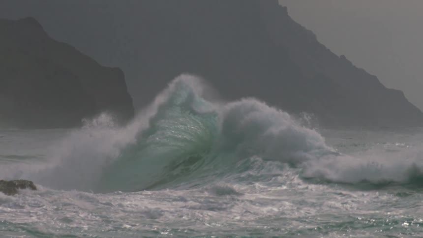 A wave is crashing against a rock in the ocean with a stretch of land in the background
