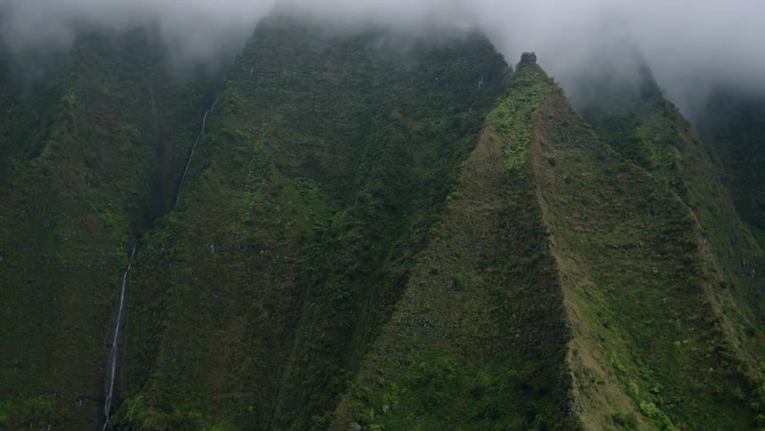 An aerial view showcases steep verdant mountain slopes and cliffs partially veiled in lowhanging clouds