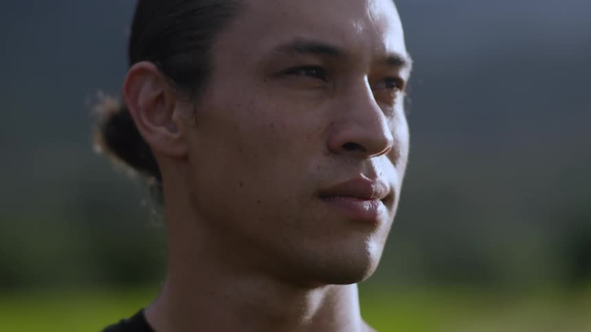 A contemplative man with tiedback hair gazes into the distance softly lit against a blurred natural background