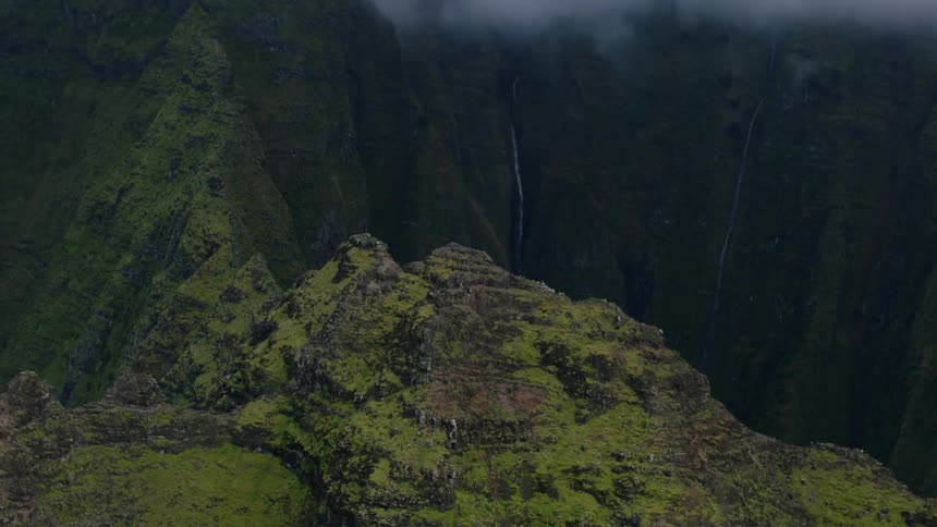 An aerial view showcases steep green mountain slopes and cliffs with a thin waterfall and lowhanging clouds
