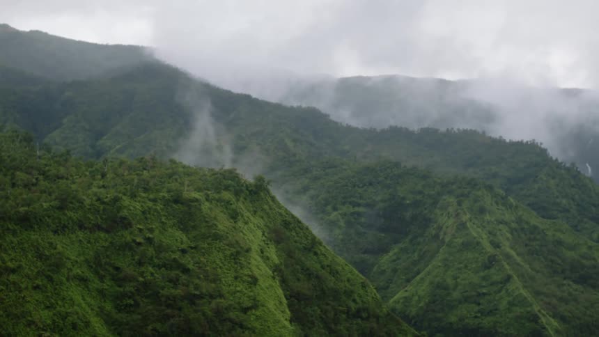 A panningright aerial view shows lush green mountain slopes and hills partially obscured by lowhanging clouds