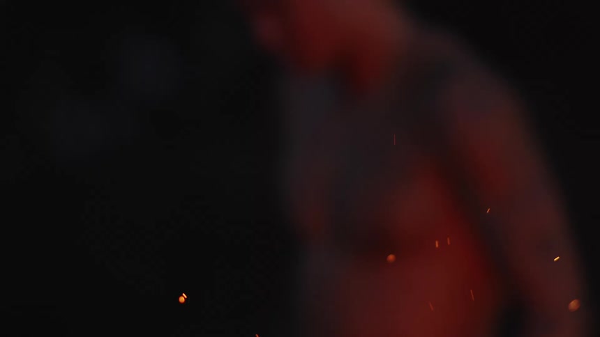 A shirtless man stands near a fire that releases embers into the air