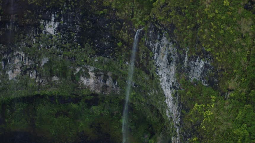 An aerial view shows a tall thin waterfall cascading down a lush green and rocky cliff face within a gorge