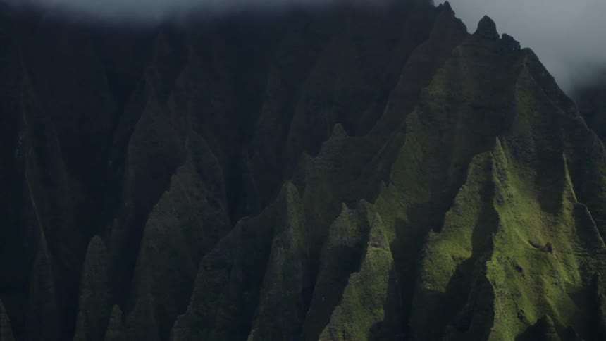 A panningright aerial view shows steep dramatic mountain cliffs with green vegetation partially shrouded in shadow