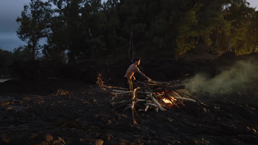 Man adds a stick to a small bonfire burning next to a grove of trees under an overcast sky