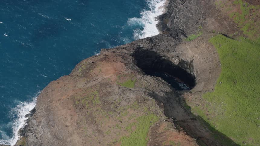 A rugged coastal cliff with a large dark cave opening near the edge with blue ocean waves crashing against the rocks below