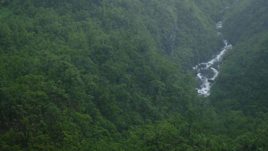 A narrow winding river cuts through a densely forested mountainous landscape under an overcast sky