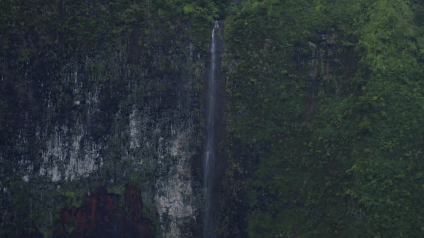 A panningdown view shows a tall waterfall cascading down a lush green cliff face ending in white wash streams at the base